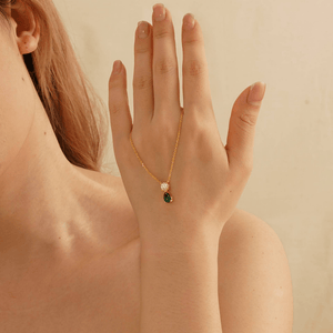 Hand wearing a gold necklace with a green gemstone on a beige background