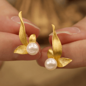Gold leaf earrings with pearls held between fingers against a blurred background