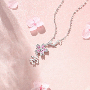 Two dainty necklaces with pink cherry blossom pendant designs, one with a gold plating and the other with a silver plating, displayed on a pink background.