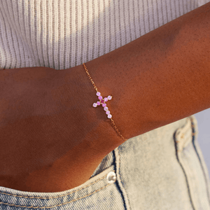 Gold bracelet with a pink and red bead on a wrist, wearing a beige top and denim jeans.