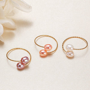 Three gold-colored bypass rings, each featuring a double pearl design in pink and white, displayed on a textured background.