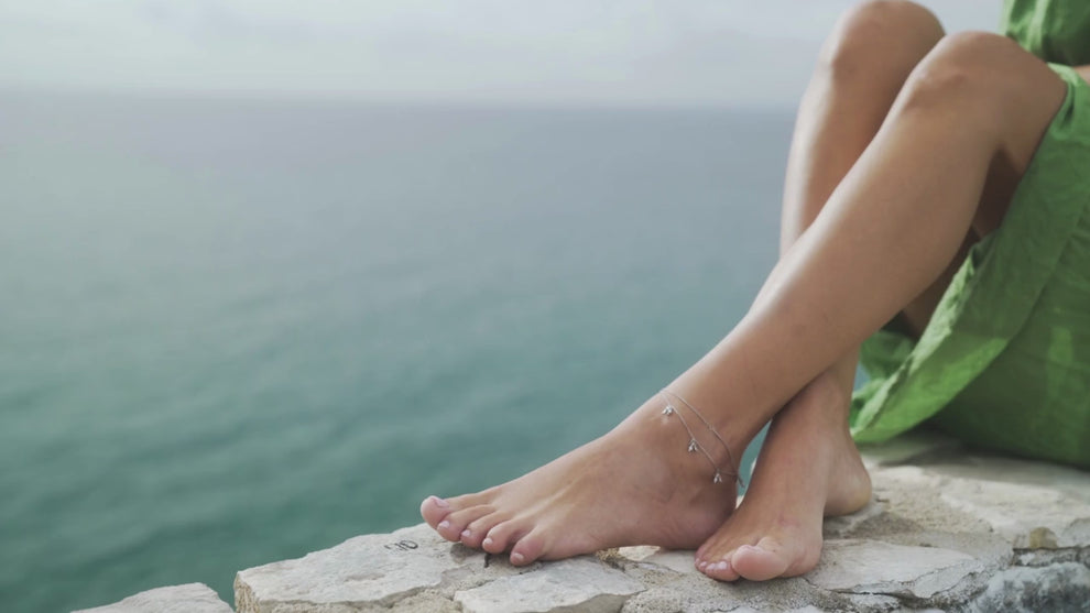 A close-up of a person sitting on a stone ledge overlooking the ocean, wearing a green dress and delicate silver anklets with small charms.