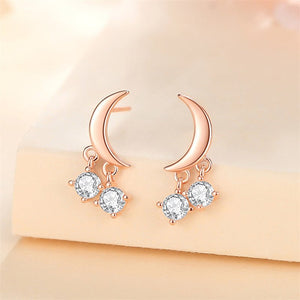 A set of three celestial-themed drop stud earrings, featuring designs resembling moons and stars, in silver and gold colors, embedded with clear stones.