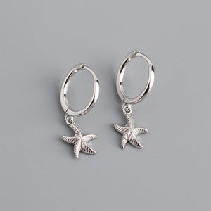 A pair of hoop earrings with starfish charms attached, plated in silver, displayed against a grey background.