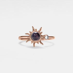 Rose gold projection ring with sun design and gemstones on a light gray background