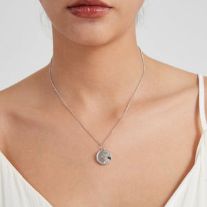 Silver necklace with a round locket featuring a sun face design, worn on a delicate chain on a model’s neck.