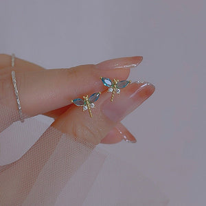 A pair of small blue dragonfly stud earrings with gold plating, displayed on a person's finger.