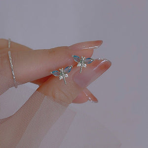 A pair of small blue dragonfly stud earrings with gold plating, displayed on a person's finger.