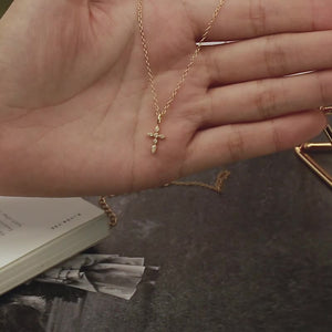 Gold necklace with a cross pendant held in a hand, with a book and paper in the background.