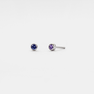 A pair of small custom birthstone stud earrings, with one earring featuring a green birthstone and the other featuring a red birthstone, both set in a solid silver plating.