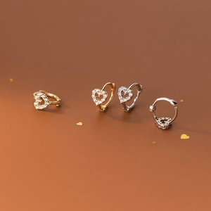 Four heart-shaped earrings in gold, rose gold, silver, and bronze on a brown background.