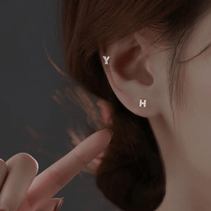 Close-up of a person wearing silver earrings with 'Y' and 'H' charms, pointing to the earrings.