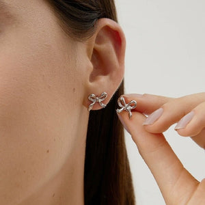 Silver bow-shaped earrings being worn and held by a hand against a neutral background