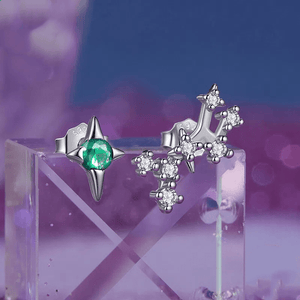 A pair of delicate zodiac stud earrings with one featuring a green cubic zirconia at the center and the other with a white gold plating and smaller white stones.