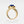 A gold-colored statement cocktail ring with a blue enamel evil eye design on a white background.