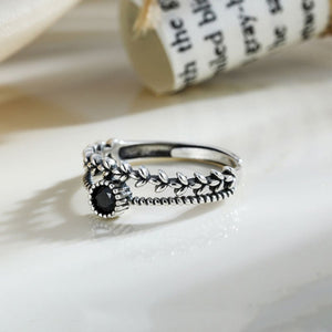 A silver stacking ring with leaf patterns and a black cubic zirconia centerpiece, displayed on a white surface with a book in the background.