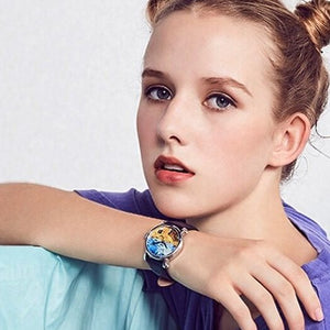 A young woman with her hair styled in two buns poses while wearing a silver-cased wristwatch. The watch face features a vibrant, artistic painting reminiscent of Van Gogh's "The Starry Night," with swirling blue and yellow colors. She is wearing a purple and turquoise top, and the image is set against a clean, light-colored background.