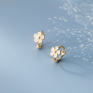 A pair of white enamel flower huggie earrings in gold plating, presented against a light blue background.