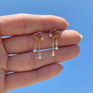 Set of colorful gold charm drop stud earrings featuring cloud and dangling chains with pastel stones, displayed on a hand against a bright blue background.