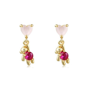 Gold drop earrings featuring heart-shaped pink stones as studs with dangling teddy bear charms holding red gemstones, set against a white background.