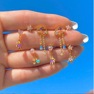 Set of colorful gold charm drop stud earrings featuring hearts, stars, bows, flowers, and dangling chains with pastel stones, displayed on a hand against a bright blue background.