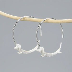 A pair of gold-colored hoop earrings with a dachshund dog figure attached, displayed against a light background.