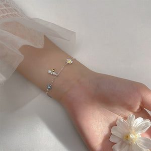 A sterling silver stacking bracelet with daisy flower charms and a small bell, displayed on a blue background with white flowers.
