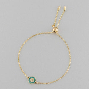 Gold bracelet with a green and gold evil eye charm on a gray background