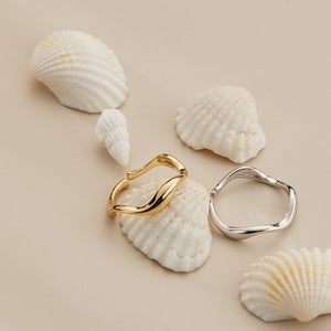 A gold and silver stacking rings with curved wave design, displayed on a beige background with seashells.