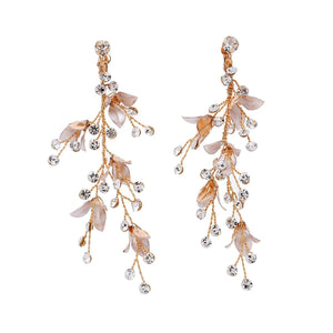 A woman wearing a white dress featuring long dangle drop earrings with gold plating, pearls, and leaf designs.