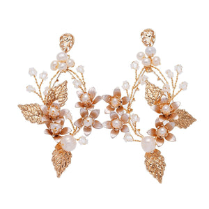 A woman wearing a white dress featuring long dangle drop earrings with gold plating, pearls, and leaf designs.