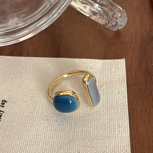 An open statement cocktail ring with gold plating, featuring a blue stone on one side and a white stone on the other, placed on a white fabric.