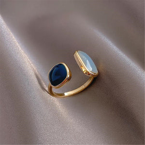An open statement cocktail ring with gold plating, featuring a blue stone on one side and a white stone on the other, placed on a silky beige fabric.