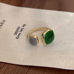 An open statement cocktail ring with gold plating, featuring a green stone on one side and a white stone on the other, placed on a white fabric.