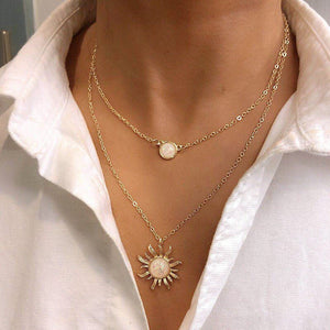 A person wearing a white shirt showcasing a gold-plated necklace with a sun charm and a moon charm, part of the necklace's double layering.