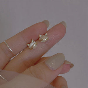 Gold cat-shaped earrings on a hand with beige nail polish against a gray background