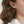 Close-up of a woman's ear featuring a small, gold tulip-shaped stud earring with a tiny sparkling stone in the center. Her wavy brown hair is tucked behind her ear, and she is wearing a white blouse.